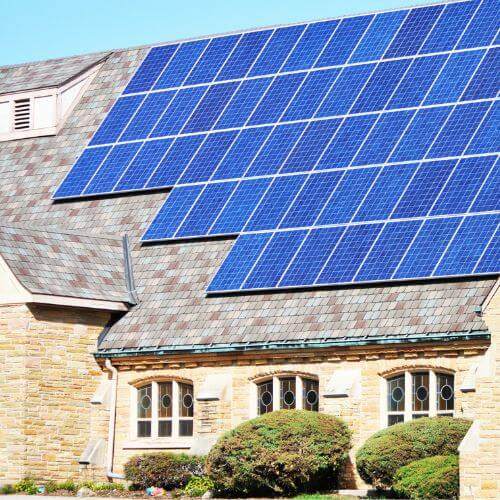 A large array of blue solar panels covers a significant portion of a sloped, shingled roof belonging to a light-colored stone building, potentially a church, demonstrating a solar energy solution suitable for the Xcel Solar Battery Storage Program.