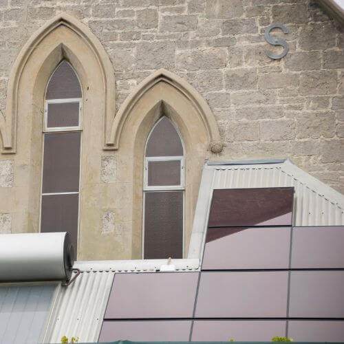 A detailed view shows dark-colored solar panels installed on a corrugated metal roof, contrasting with the light stone wall of an older building featuring arched windows and a decorative "S" emblem, suggesting a potential site for an Xcel Solar Battery Storage Program.