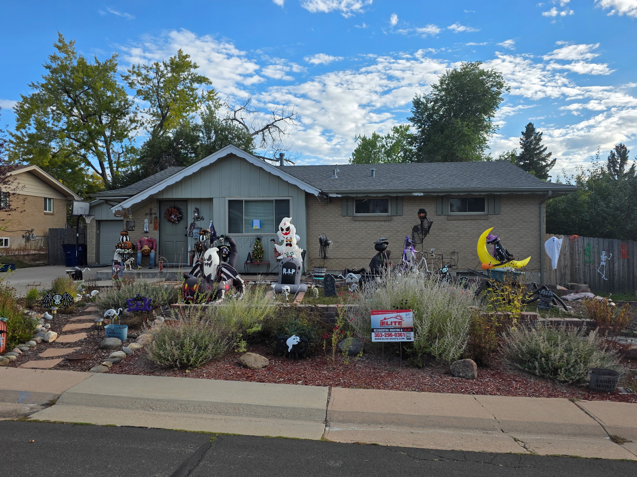 A residential, single-story ranch-style house with a recently replaced, dark gray shingle roof (possibly donated by Elite Roofing & Solar, as indicated by a yard sign). The house is beige and has a yard decorated heavily for Halloween, with various skeletons and spooky figures visible in the foreground. A red and white sign for Elite Roofing & Solar is displayed in the landscaped front yard, suggesting their work on the roof.