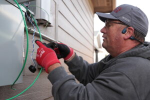 Elite employee wearing cap and red gloves using a power tool to connect electrical wiring to a wallโmounted solar system unit in Denver