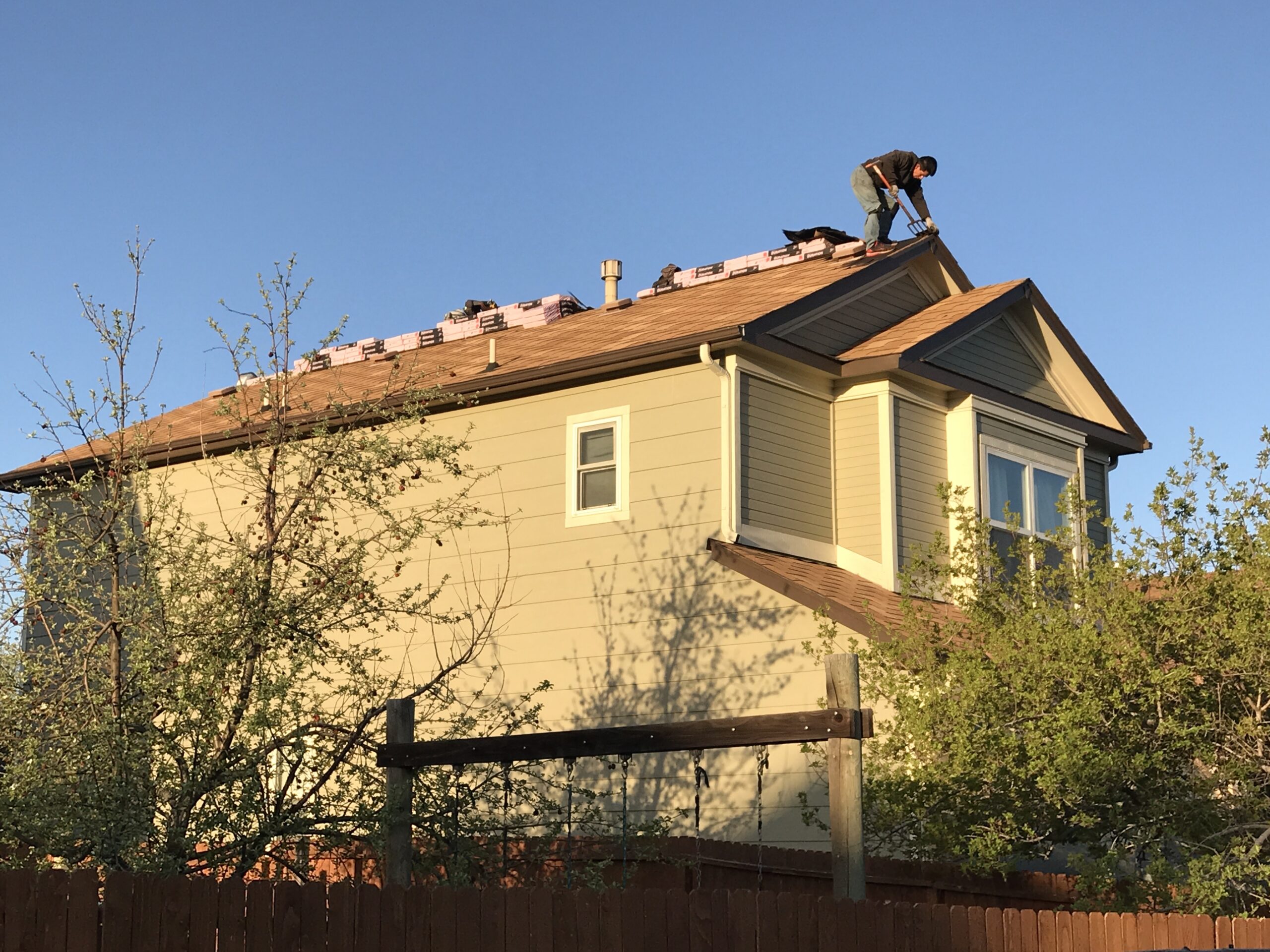A construction worker uses a tear-off tool on the ridge of a two-story residential home with new shingle bundles laid out. Roof replacement in Denver by Elite Roofing and Solar