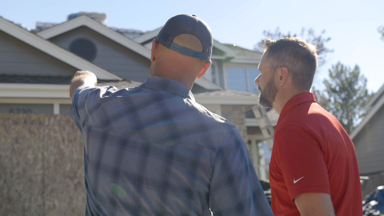 A roofing contractor in a red polo shirt discussing a residential roofing project with a homeowner who is pointing toward his house gables. Roof replacement in Denver by Elite Roofing and Solar