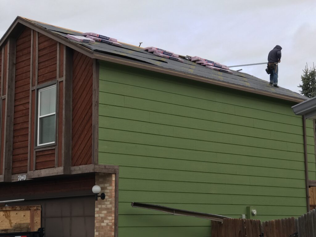 A construction worker stands on a residential roof preparing materials for a roof replacement, with bundles of pink shingles resting on the dark underlayment. Roof replacement in Denver by Elite Roofing and Solar