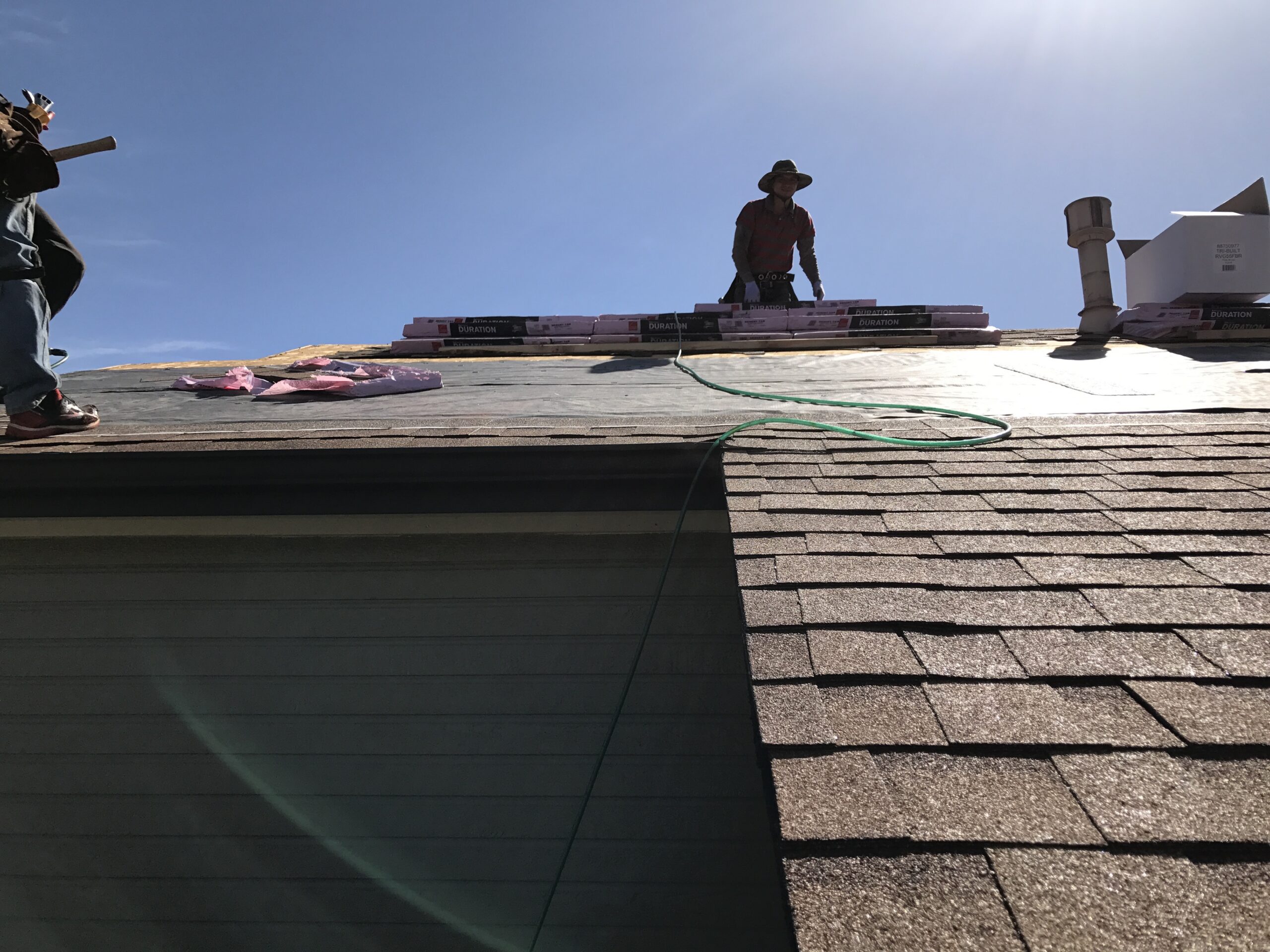 A roofer in a sun hat works near stacked shingle bundles along the ridge to calculate precise roof square measurements. Roof square measurements in Denver by Elite Roofing and Solar