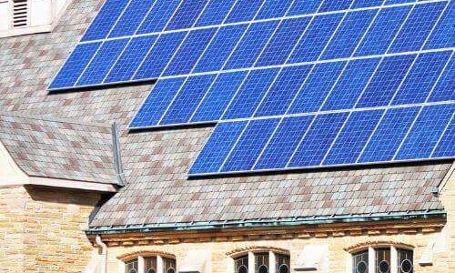 A large array of blue solar panels covers a significant portion of a sloped, shingled roof belonging to a light-colored stone building, potentially a church, demonstrating a solar energy solution suitable for the Xcel Solar Battery Storage Program.
